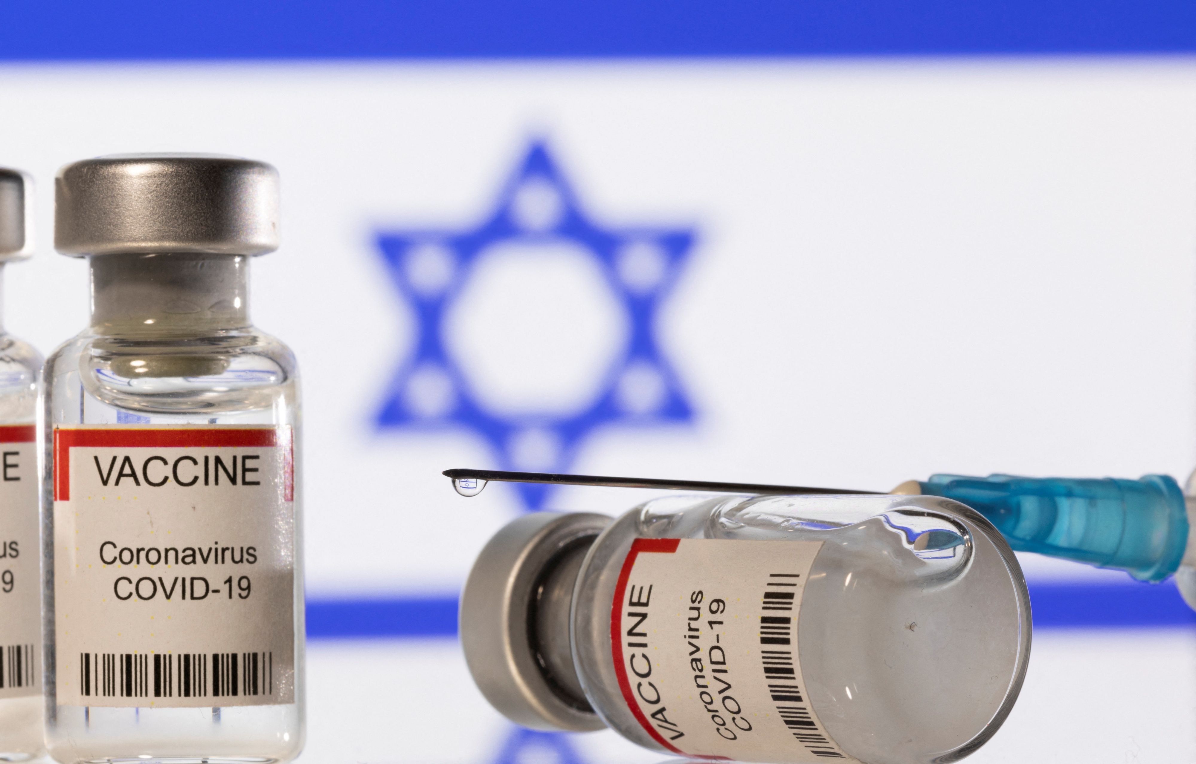 Illustration shows vials labelled "VACCINE Coronavirus COVID-19" and a syringe in front of a displayed flag of Israel