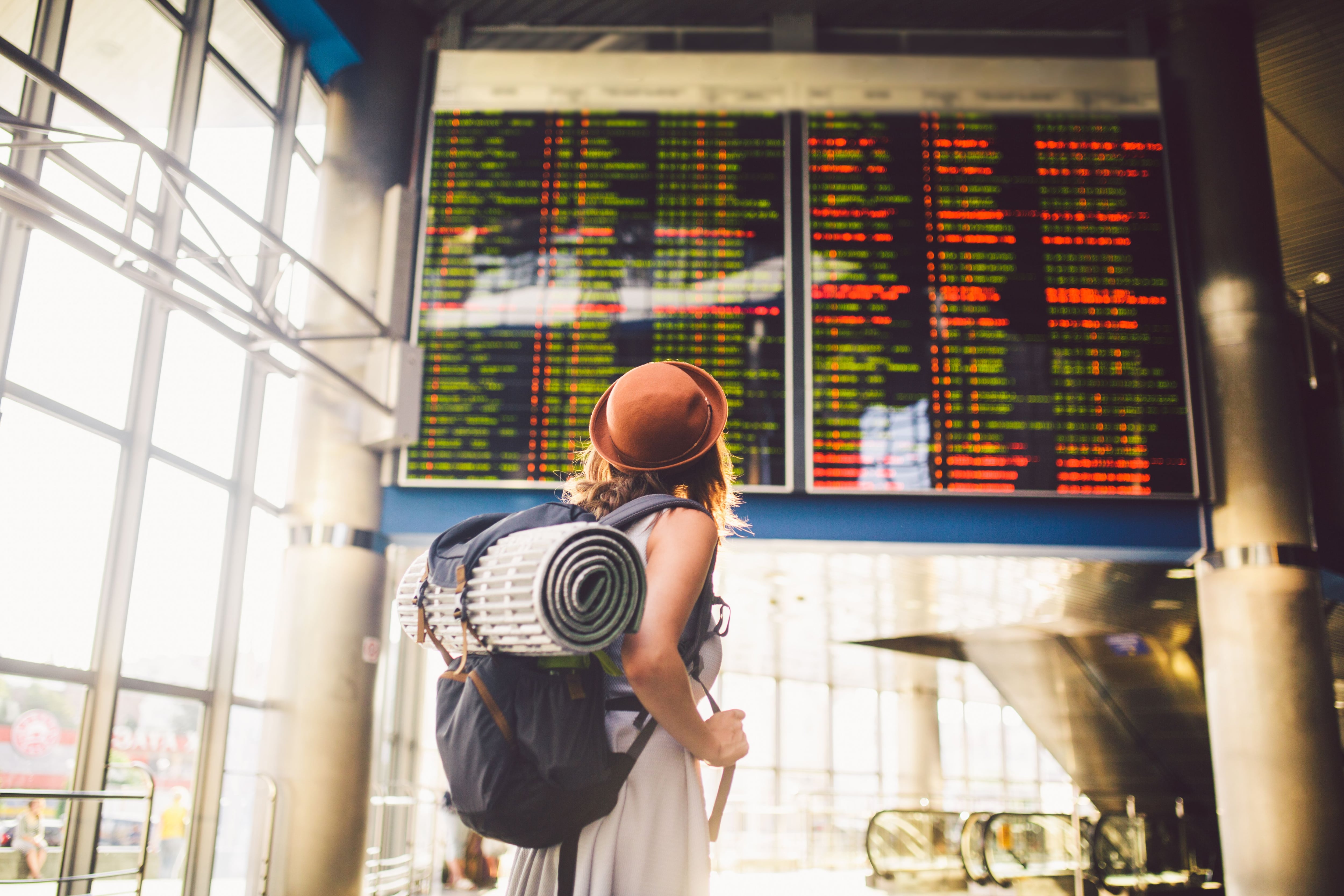 Theme travel public transport. young woman standing with back in dress and hat behind backpack and camping equipment for sleeping, insulating mat looks schedule on scoreboard airport station